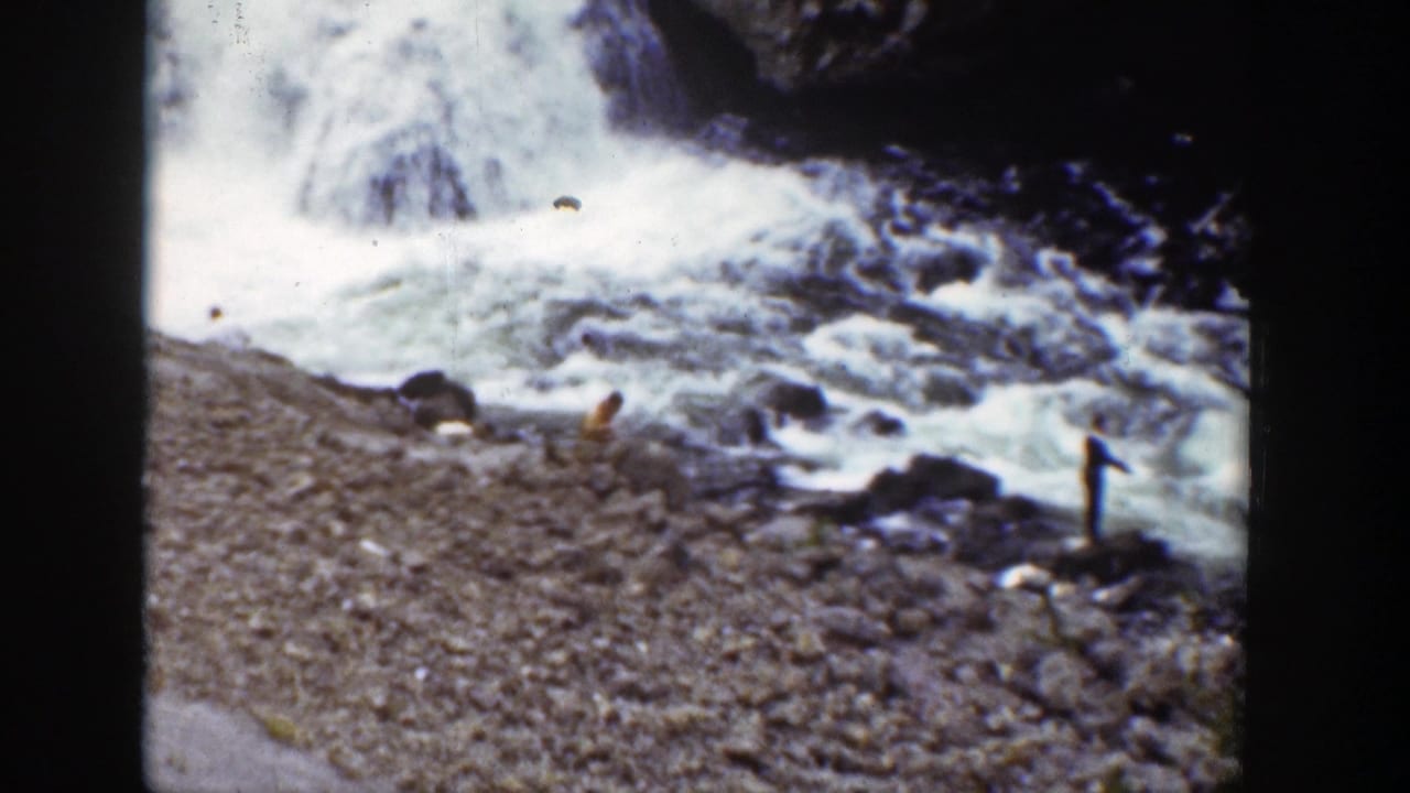 Rushing water beside a rocky bank with dark film borders and a small distant figure-like shape near the stream.