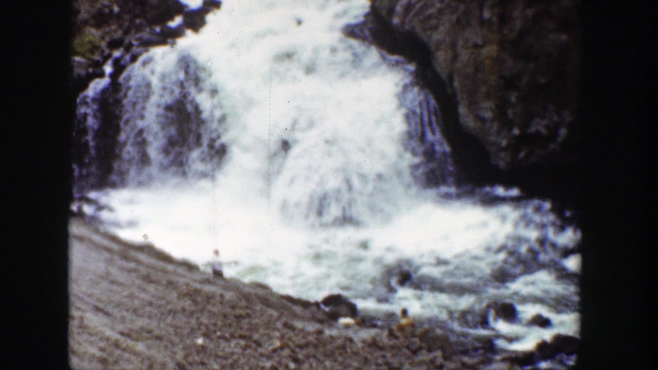 Waterfall crashing into a rocky river with small people on the gravel bank.