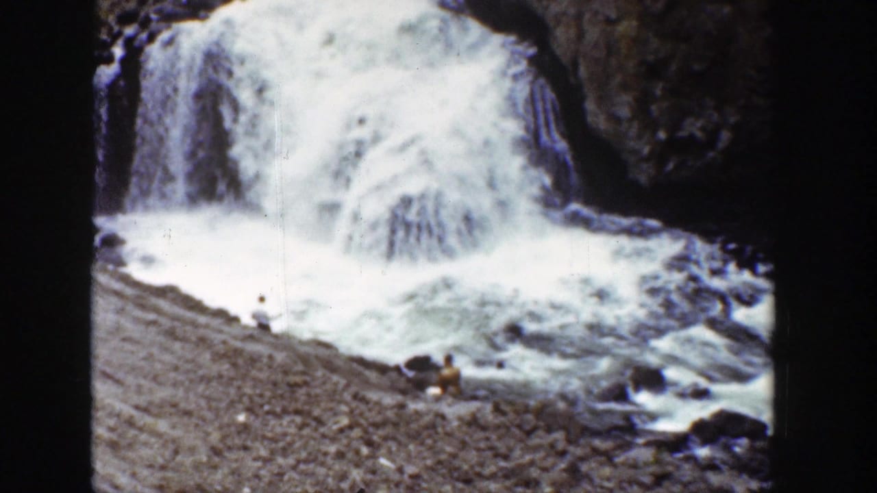 1939 Wyoming home-movie frame showing white water pouring over dark stone into a rocky river with distant visitors nearby.