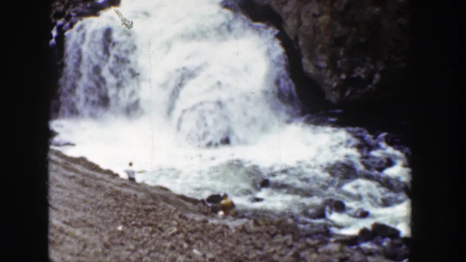 Soft 8mm frame from Wyoming in 1939 with cascading white water, dark rocks, a sloped gravel bank, and tiny people at the river edge.