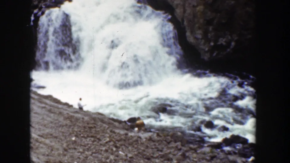 1939 Wyoming 8mm still of a bright waterfall, foamy current, dark stone, and small people near boulders on the riverbank.