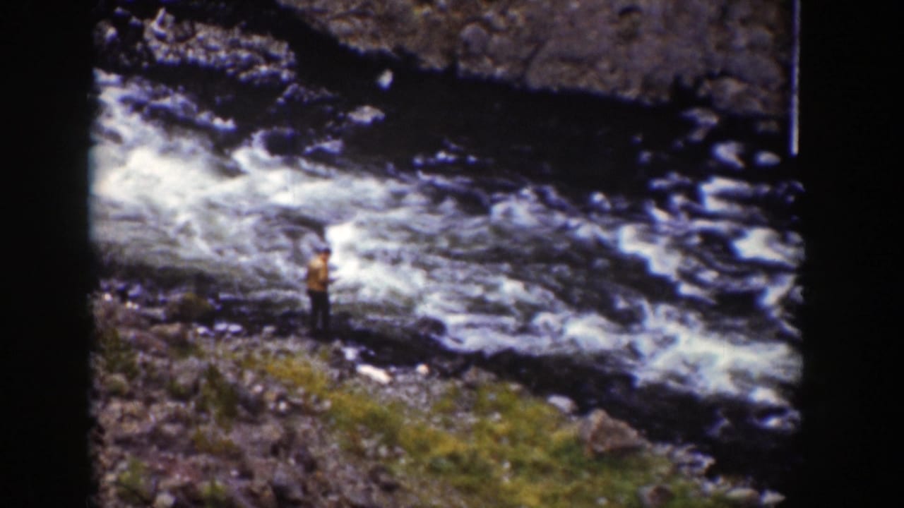Man standing near a fast river with white water over dark rocks.