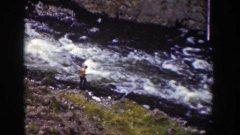 1939 Wyoming home-movie frame of a man standing on uneven ground near turbulent river water and low grass.
