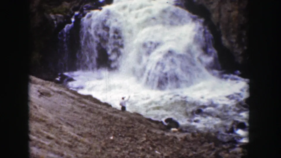1939 Wyoming frame with a waterfall crashing into white spray and a man standing on the rocky bank nearby.
