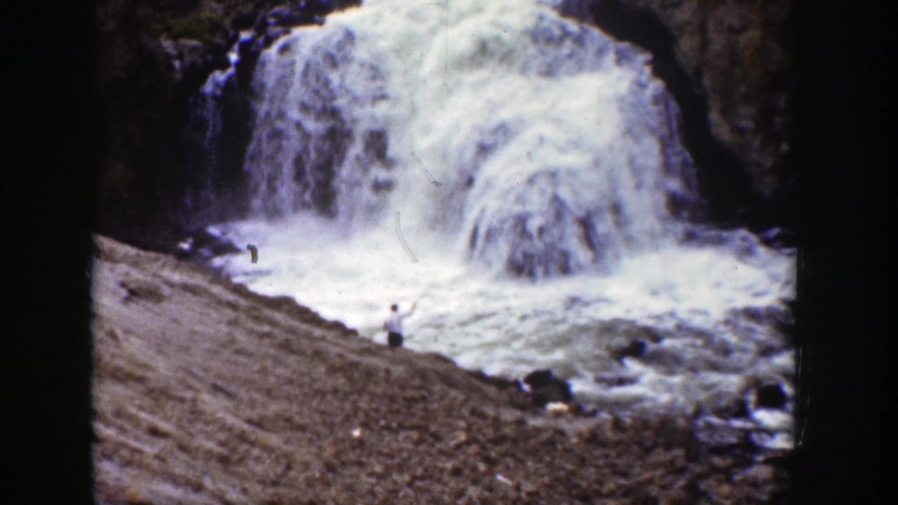 A small figure stands on a rocky bank below a broad waterfall and foaming river.