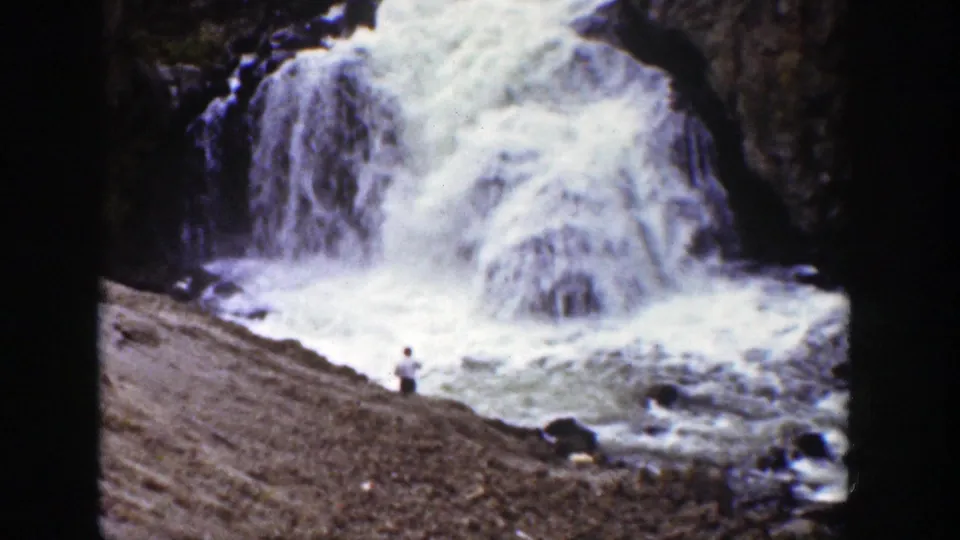 1939 Wyoming home-movie frame of a distant person watching a rushing waterfall from a rocky shoreline.
