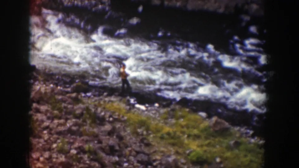 The home-movie frame shows a person standing on uneven rocks near a rushing Wyoming river, with grass in the foreground.