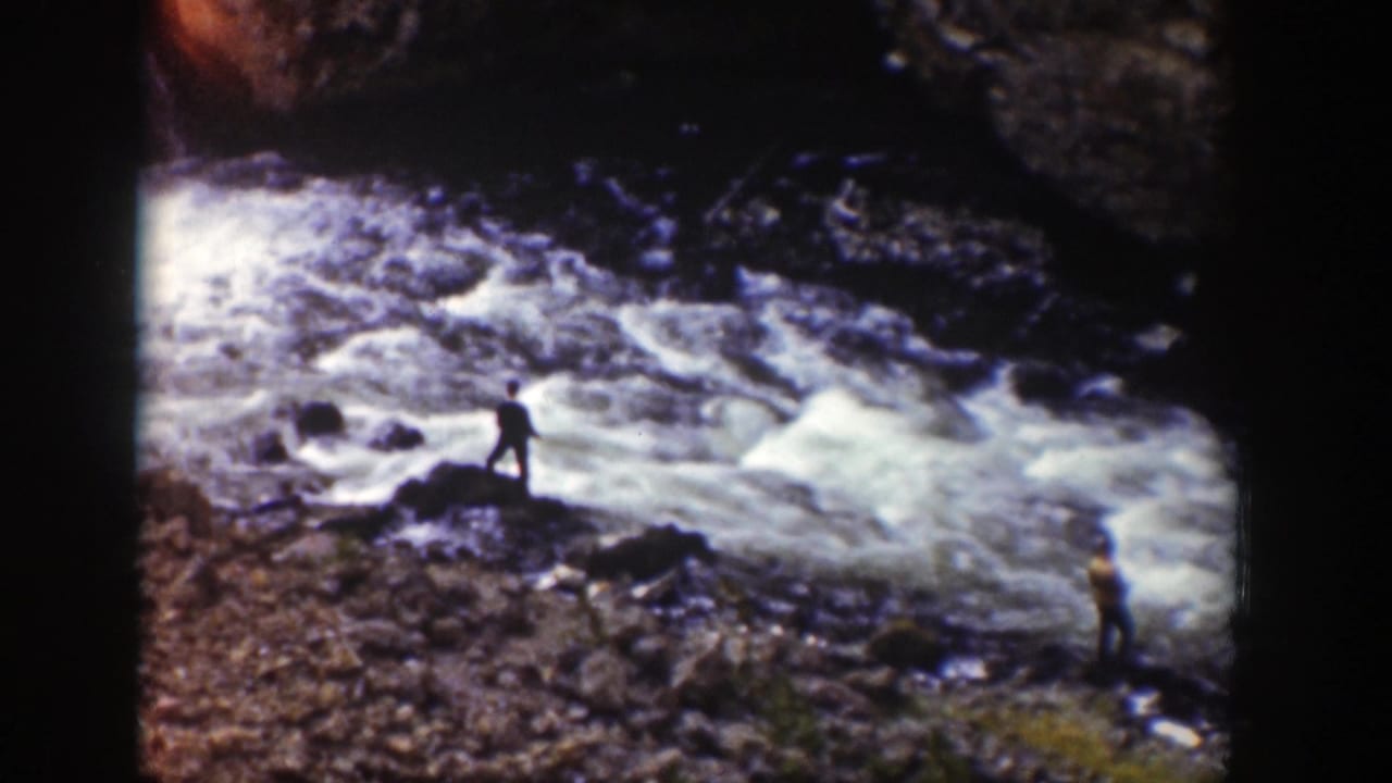 1939 Wyoming home-movie frame with a distant person on a rocky riverbank beside rushing rapids.