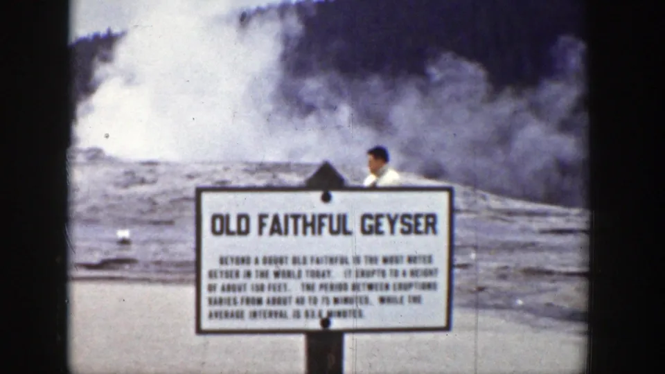 A visitor appears partly behind the Old Faithful Geyser sign as steam rises across the Yellowstone geyser basin.