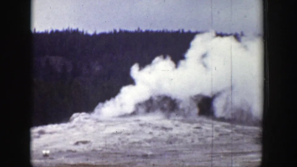 1939 Wyoming 8mm frame showing a geyser eruption beginning or receding near a conifer forest under a pale sky.