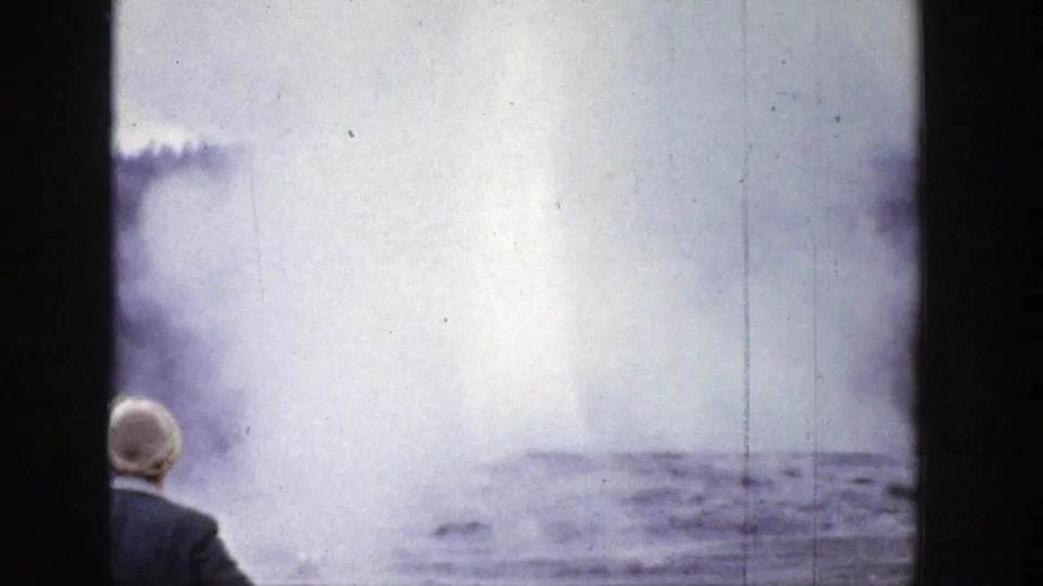 1939 Wyoming 8mm frame showing a distant person from behind watching geyser steam rise over a rocky geothermal area.