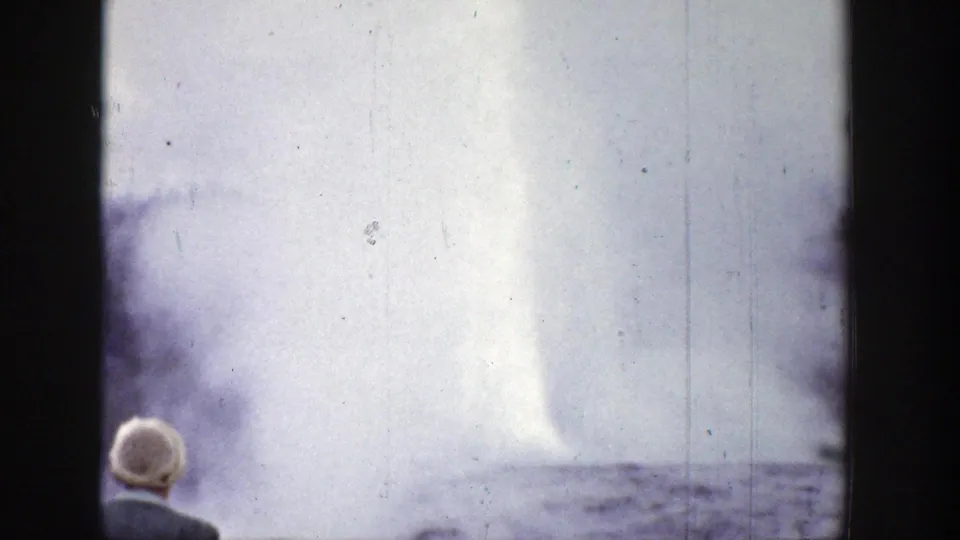 1939 Wyoming 8mm frame showing a geyser plume rising in bright sky while a white-haired spectator stands at lower left.
