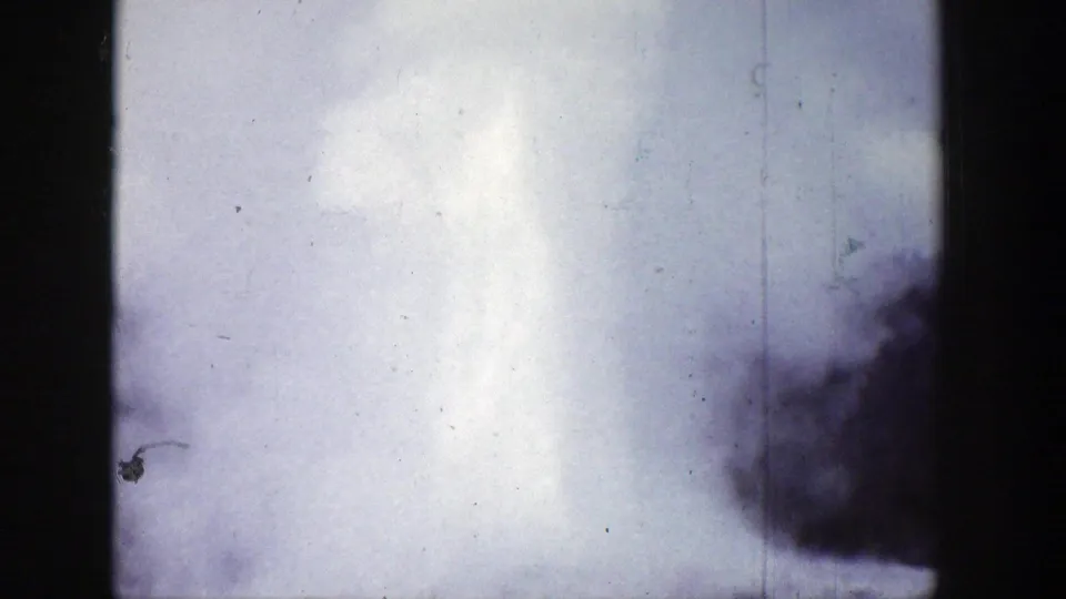 1939 Wyoming 8mm frame with geyser spray drifting upward, dark film edge at left, and blurred foliage at lower right.