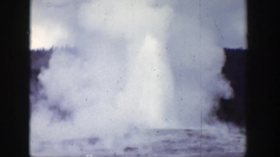 1939 Wyoming 8mm frame showing a geyser erupting in a pale plume of steam and water near dark trees.
