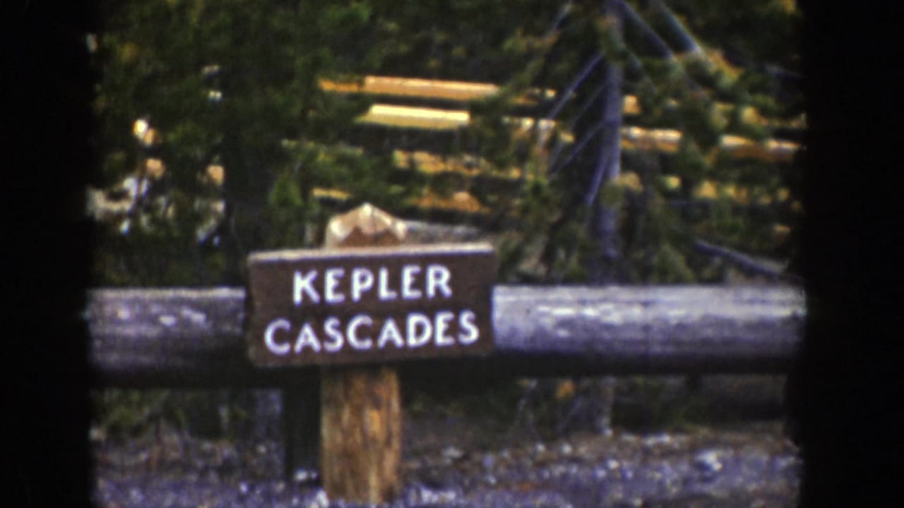 1939 Wyoming home-movie still with the Kepler Cascades marker framed by a log rail and surrounding forest.