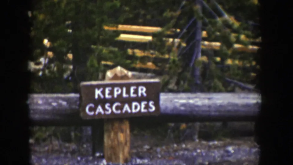 1939 Wyoming home-movie still with the Kepler Cascades marker framed by a log rail and surrounding forest.