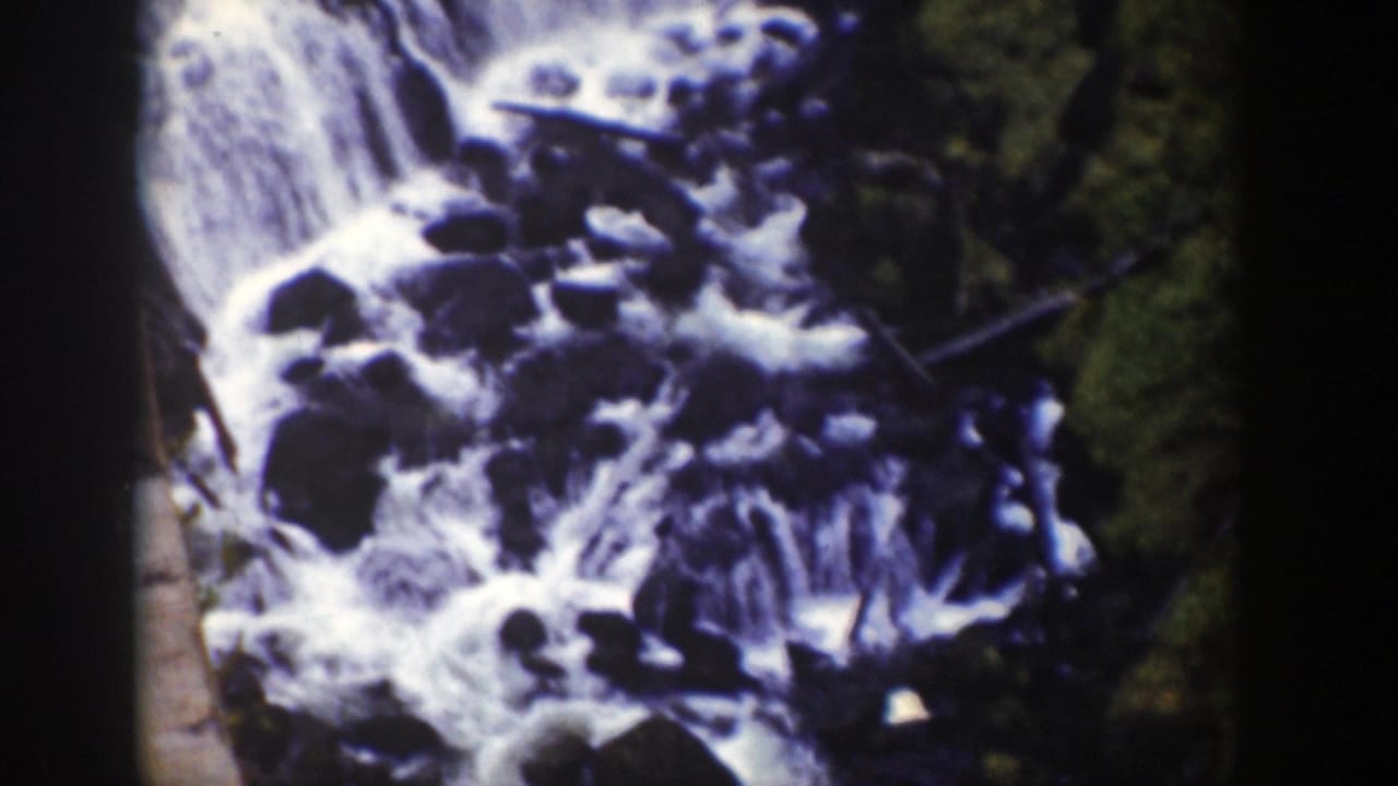 Cascading water flows around large rocks with trees and a dark foreground strip.