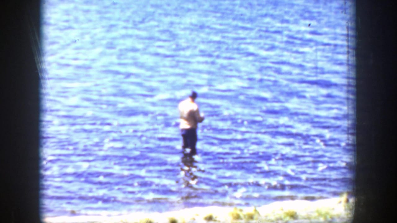 A distant adult man stands in a blue Wyoming lake, wearing a light shirt and dark pants while facing the water.