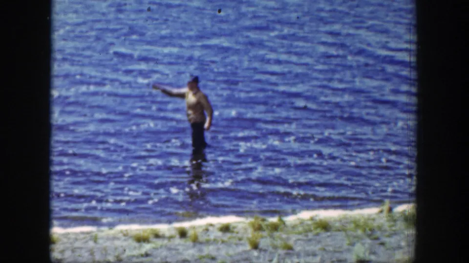 A man wades near the lake shore in Wyoming, extending one arm as though casting or handling a fishing line.