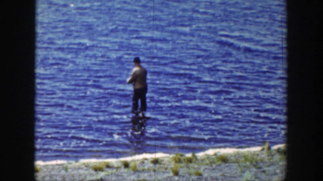 Man in a hat standing in a blue lake near a narrow shoreline.