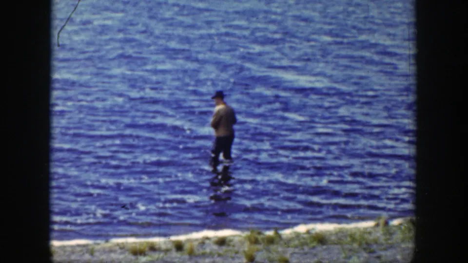 1939 Wyoming home-movie frame of a hat-wearing adult man fishing from shallow blue lake water.