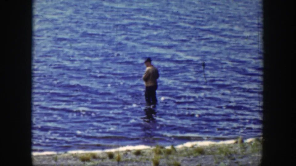 Adult fisherman stands in vivid blue Wyoming lake water, with a fishing line visible in the 1939 8mm frame.
