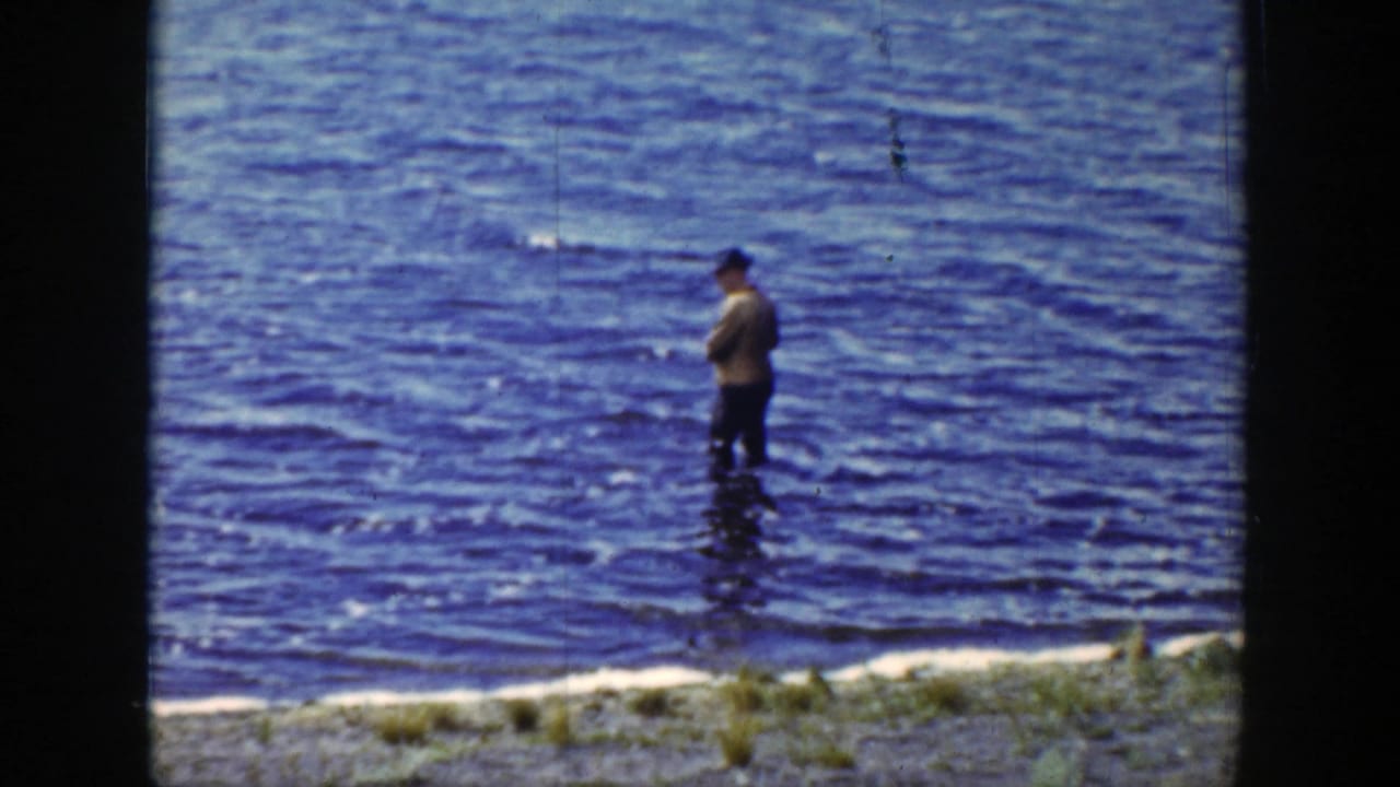 Man in a hat standing in shallow river water near a grassy bank.