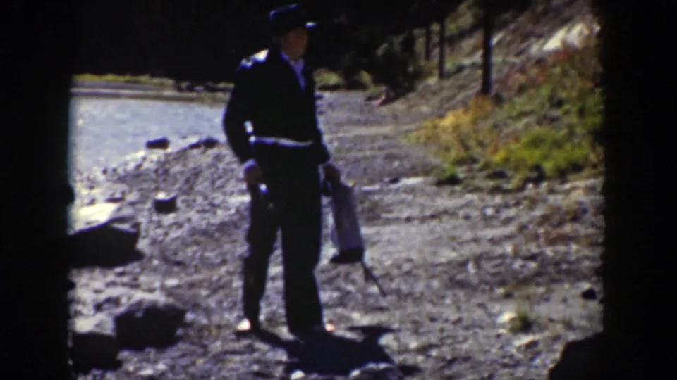 A man in a dark military-style uniform stands on a dirt path near water and trees, holding a sword and bag.