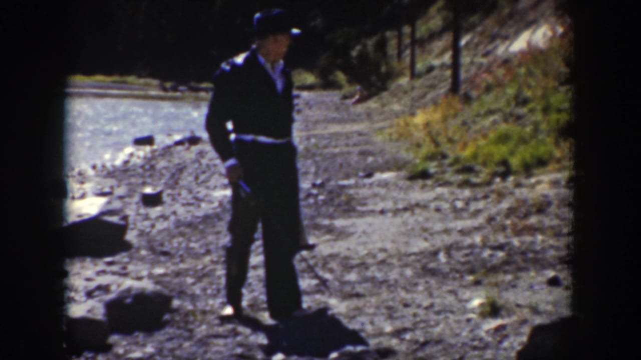 Man in dark clothing and black hat standing on a rocky stream bank.
