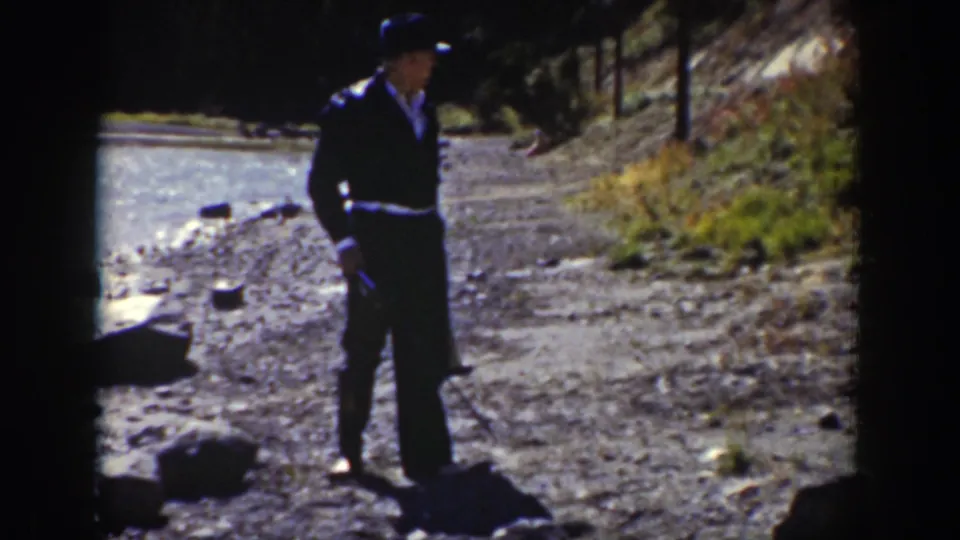 In 1939 Wyoming footage, a man in a dark suit and black hat stands near a sunlit rocky stream bank.
