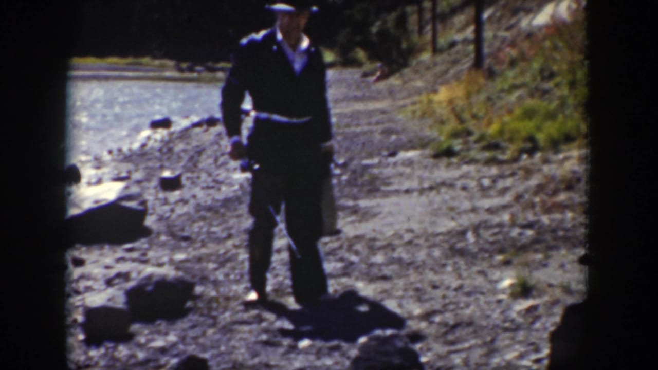 The man walks along the rocky stream edge in Wyoming, wearing dark clothing and carrying small items.