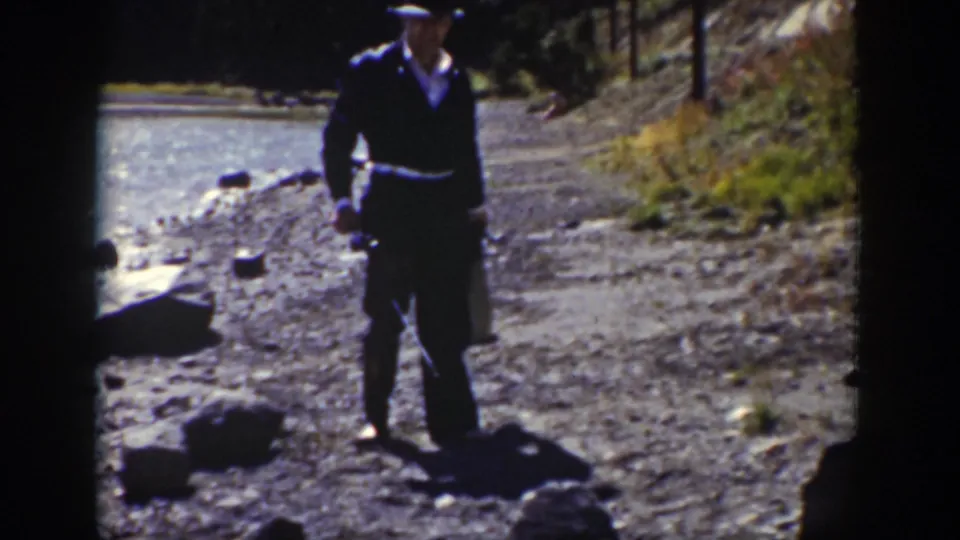The man walks along the rocky stream edge in Wyoming, wearing dark clothing and carrying small items.