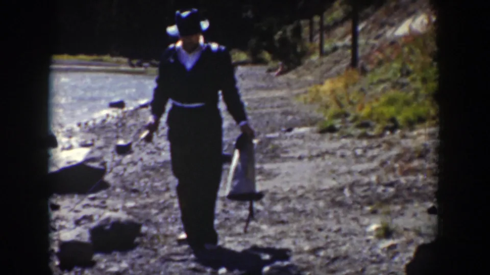 A man in dark 1930s-style clothing approaches the camera on a rocky Wyoming stream bank with a bag and long object.