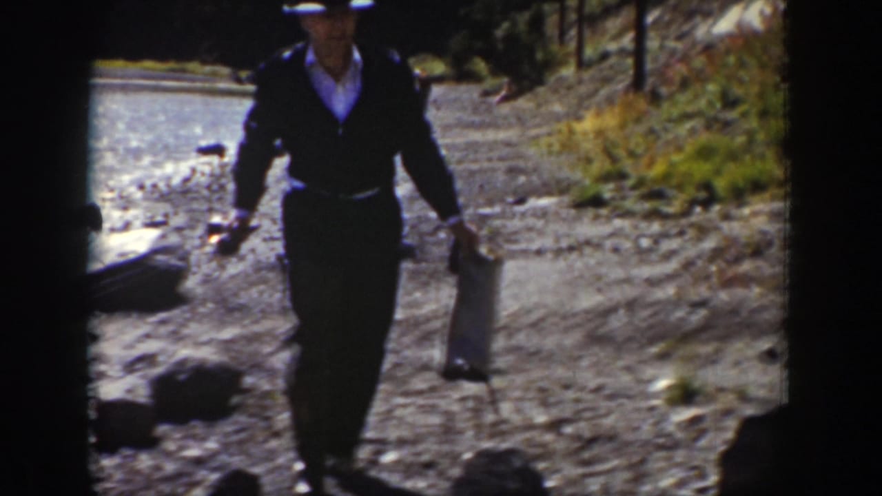 Man in a dark hat walks along a rocky shoreline carrying fishing gear.