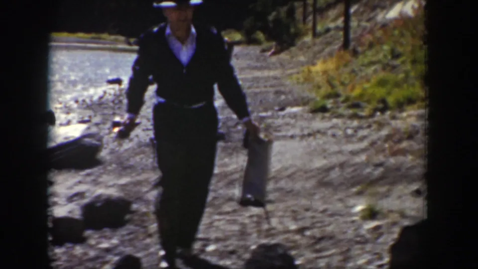 In Wyoming 1939 home-movie footage, a man in dark clothing walks on a rocky shore with fishing gear near the water.