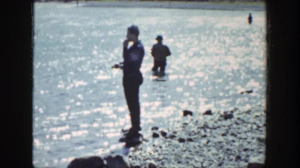 A man stands on the rocky Wyoming shoreline in 1939 footage, with another man wading in the reflective water behind him.
