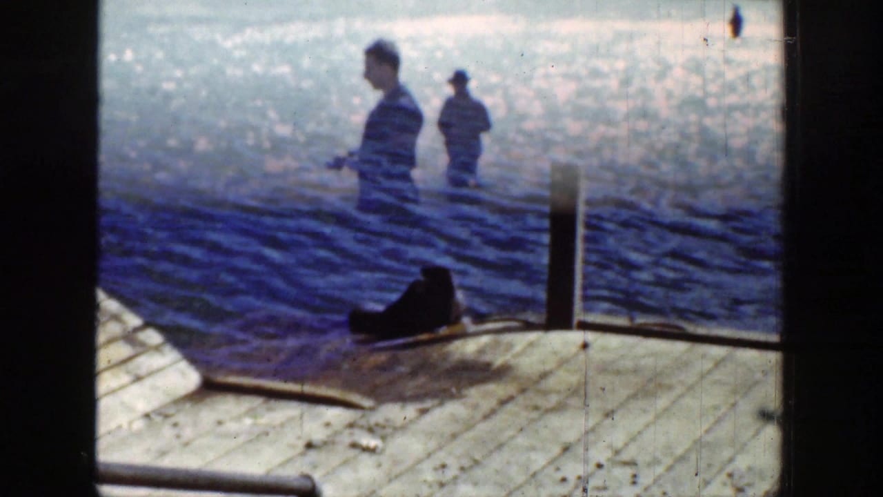 Man fishing beside rippled water with another figure farther back and a dark animal near a wooden dock.