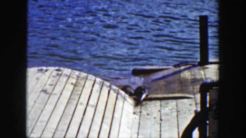 1939 Wyoming home-movie frame showing an otter at the edge of a weathered wooden dock near lake water and a boat.