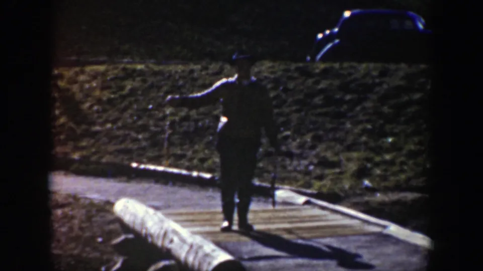 A woman in dark period clothing stands near a roadside log barrier, with a vintage car visible in the background in Wyoming.