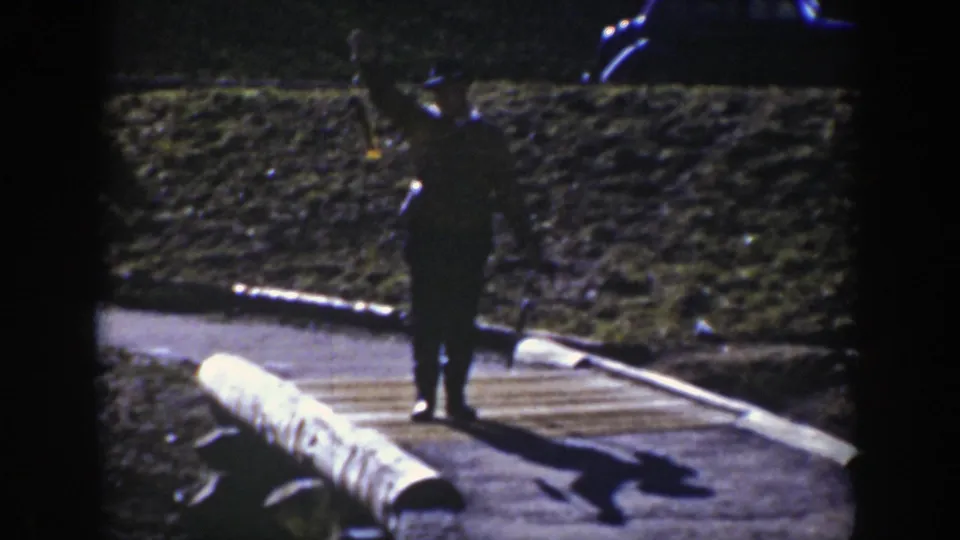 A man in a dark uniform or official-looking clothing stands on a Wyoming road near a log barrier and vintage car.