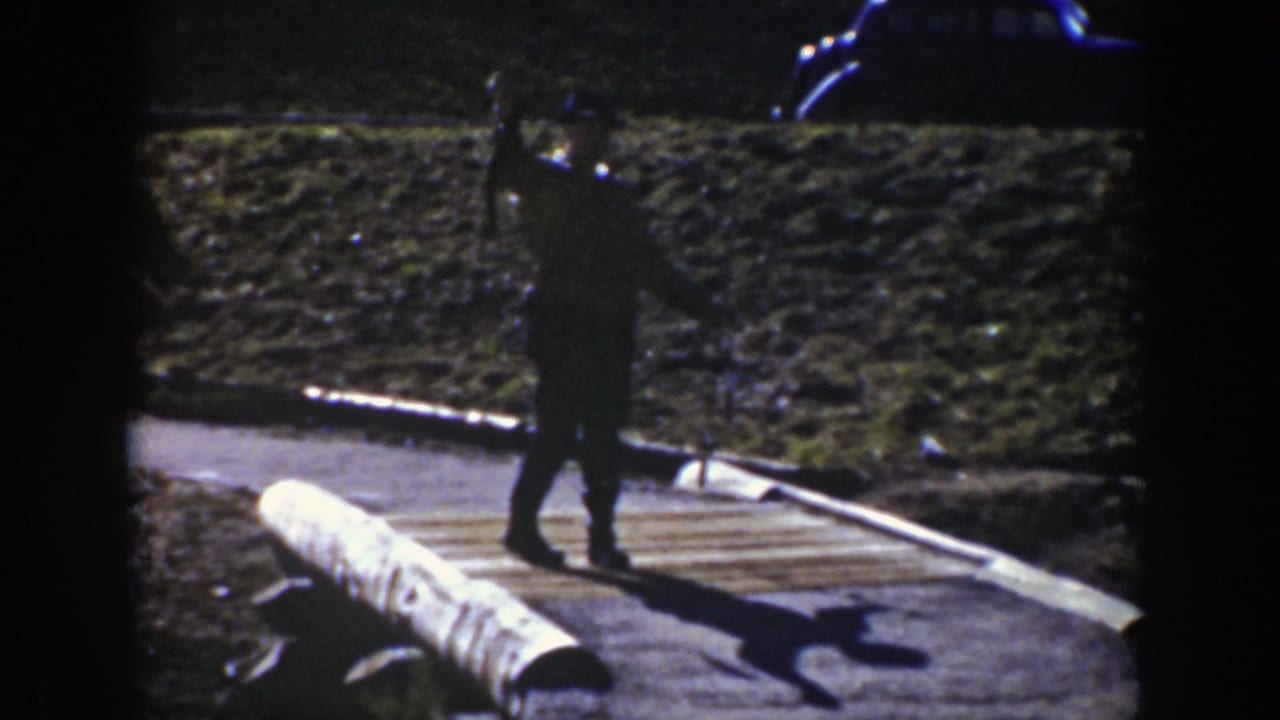 A dark-clad person stands on a pathway near rocky ground and a large pipe-like object.