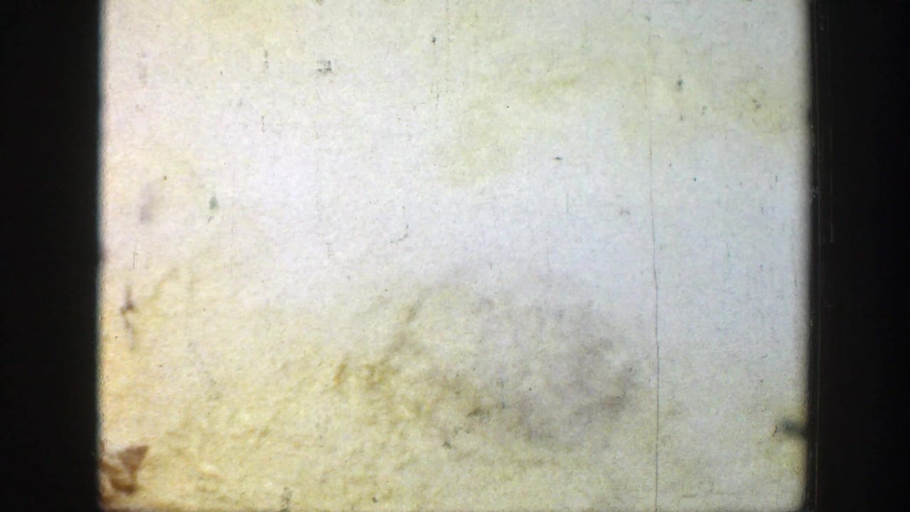 A pale, yellow-tinted 8mm frame with scratches, dust, and a dark vertical gate edge obscuring scene detail.