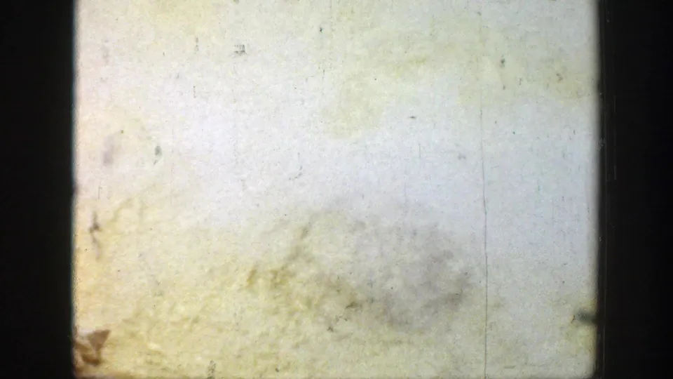 A pale, yellow-tinted 8mm frame with scratches, dust, and a dark vertical gate edge obscuring scene detail.