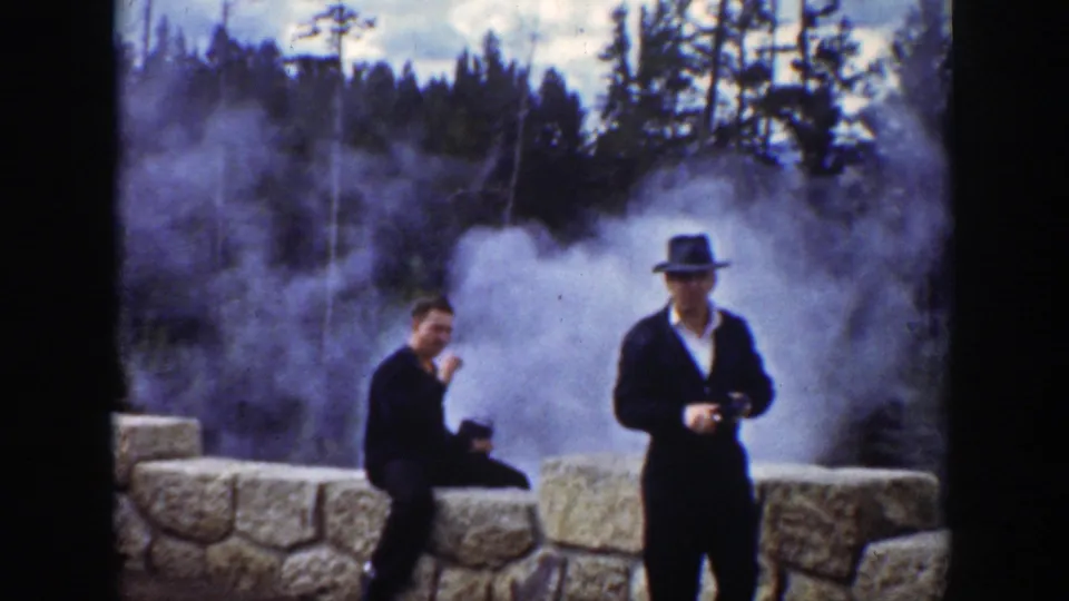 Two men in suits and brimmed hats appear at a Wyoming geothermal overlook as steam rises behind a stone wall and trees.