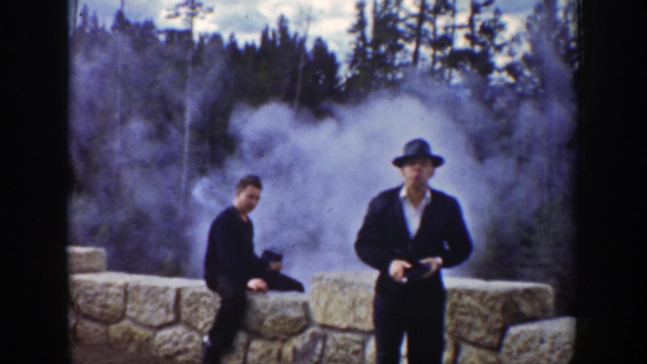 Two men near a stone barrier with geyser steam and trees behind them.
