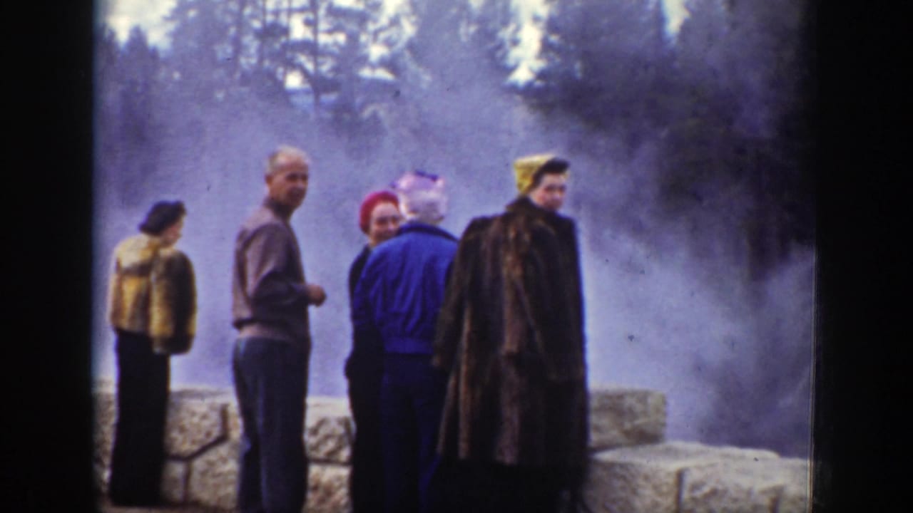 Tourists in coats, hats, and scarves stand beside a stone barrier at a Wyoming geothermal site, with dense steam in the background.
