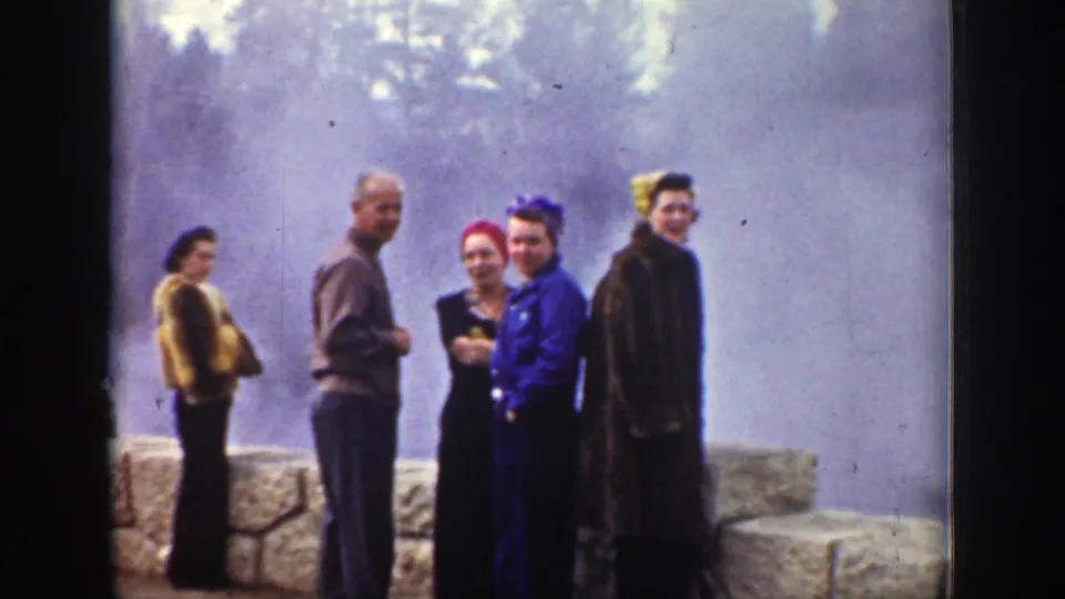 1939 Wyoming frame of several tourists posing near a tan stone barrier, with purple-tinged geyser steam obscuring the trees behind them.