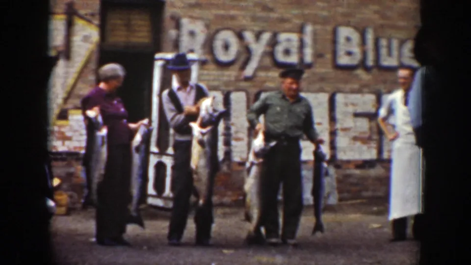 In Wyoming 1939 footage, three adults display large fish outside a brick building with a faded Royal Blue wall sign.