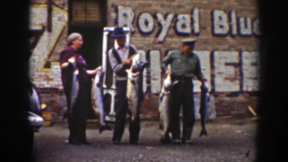 Three adults in 1939 Wyoming home-movie footage hold large fish near a brick wall, with part of a parked car at frame edge.