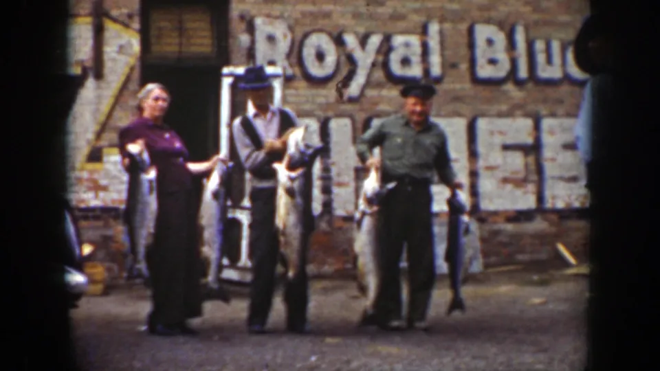 A woman in a purple dress and two men hold large fish in front of a faded Royal Blue sign in Wyoming 1939 footage.